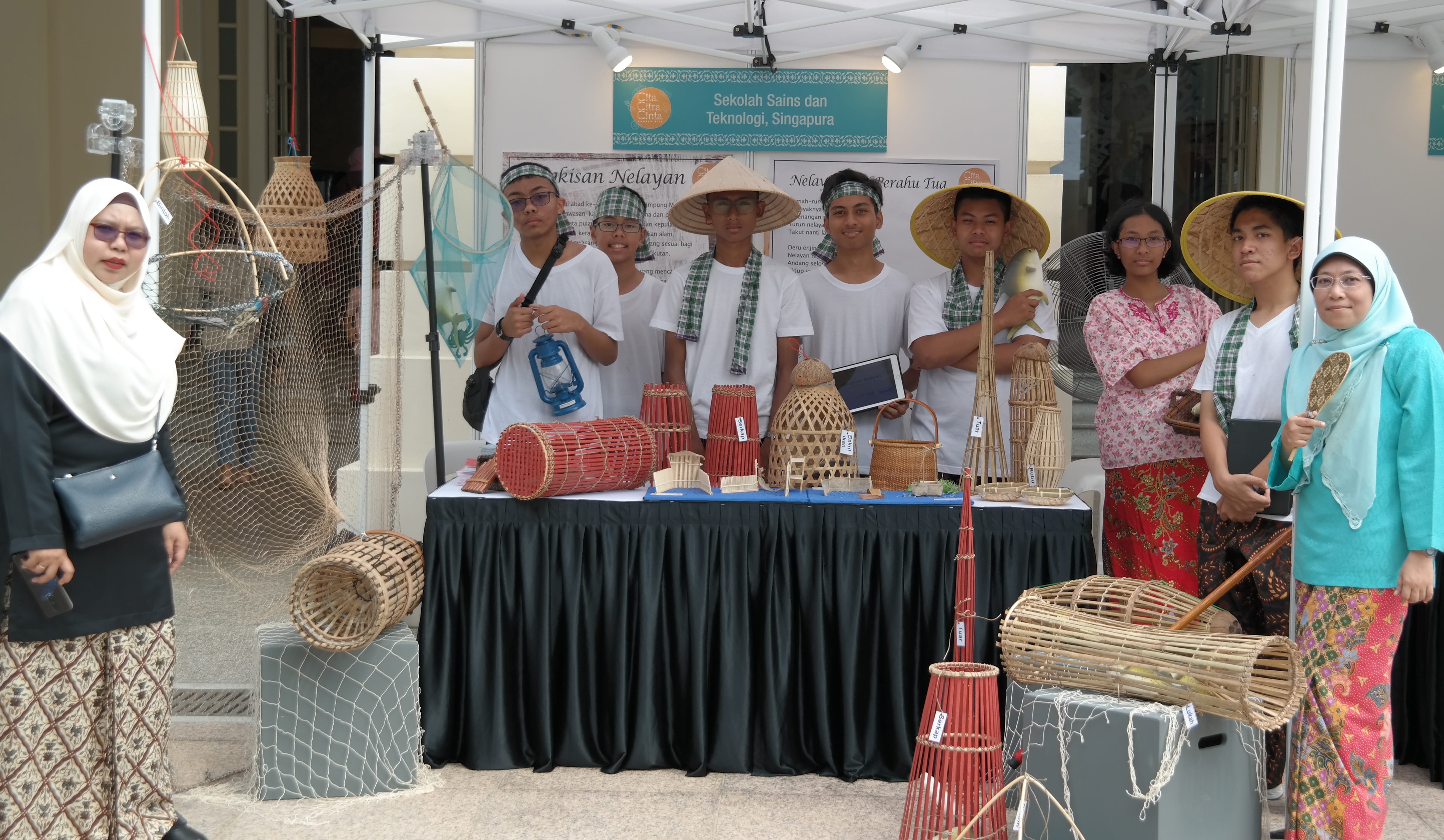 Students in traditional attire stand at a Singapore school exhibition booth displaying woven bamboo fish traps and nets.